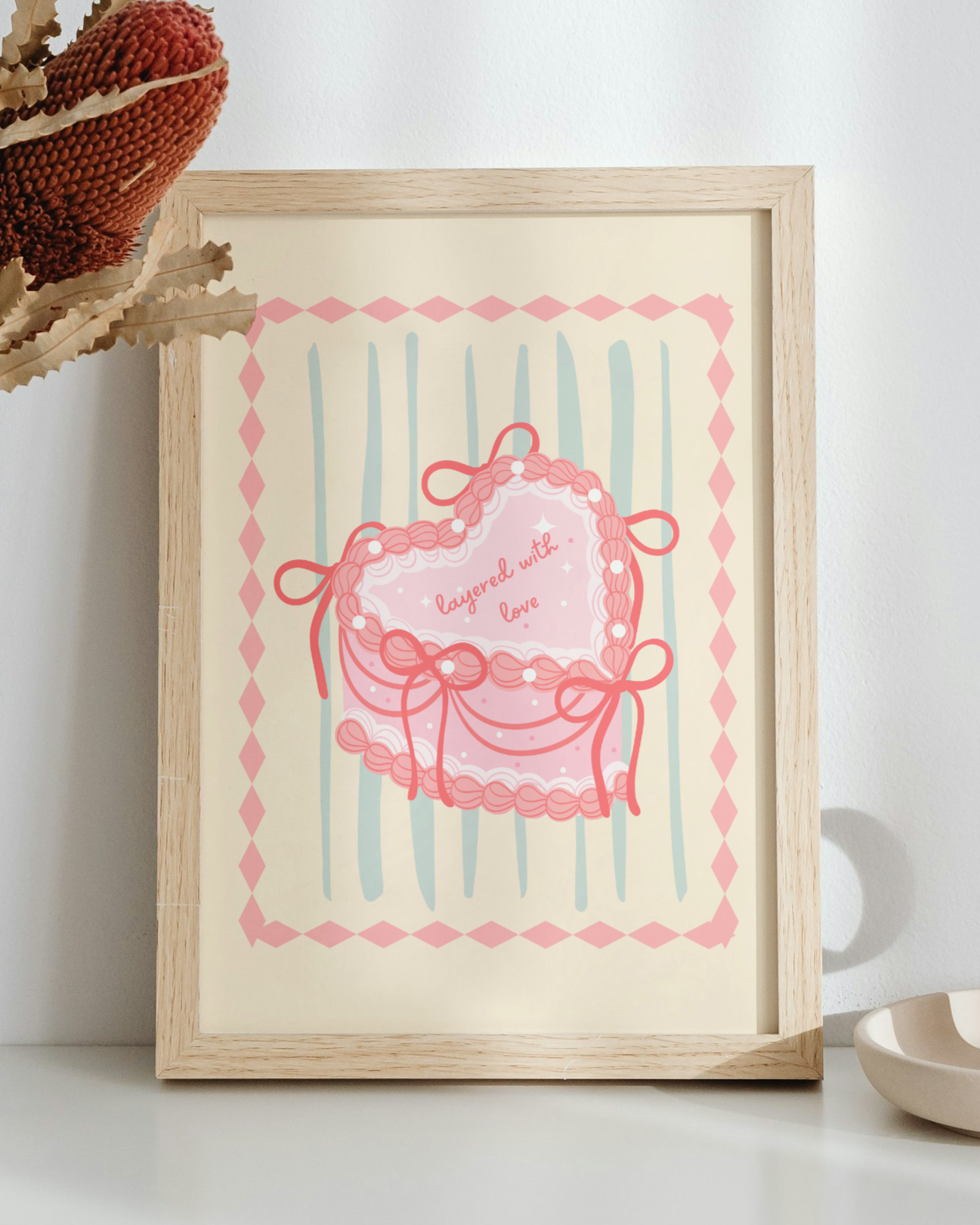 Decorative setup with a vase of dried plants, framed artwork, and a candle on a white surface. The print is of a vintage heart cake with the text "layered with love". The print is Pink, cream and seafoam green pastel.